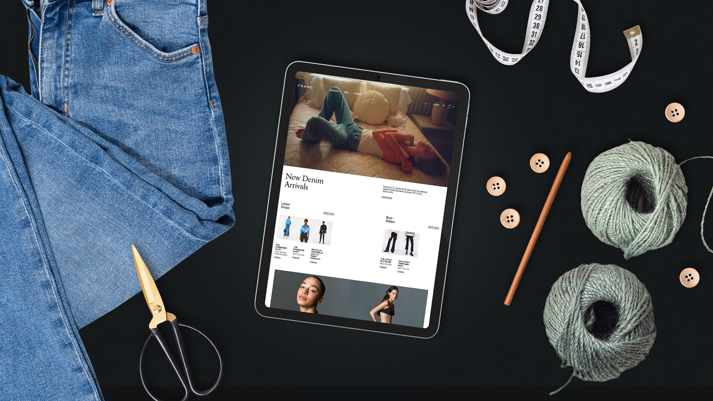 Flatylay of FRAME website on an iPad surrounded by a pair of jeans, threads, buttons, scissors, and a tape measure.