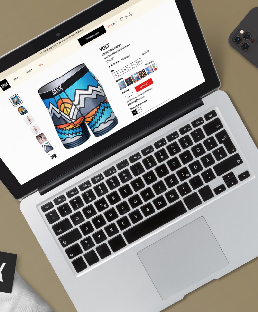 A laptop showing a product page on the SAXX website with select personal items including a wallet and a cell phone on the desktop beside the laptop.